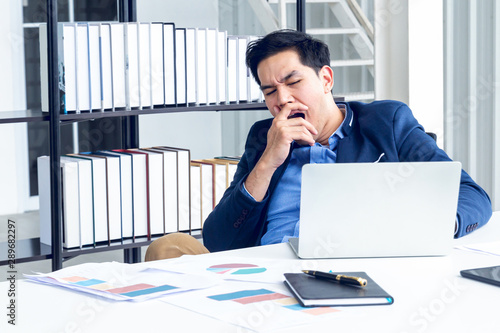 A young businessman sitting in a modern office. He has a feel sleepy because  hard work so tired weary fatigued and exhausted. On his table have a computer laptop tablet pen paper graph.