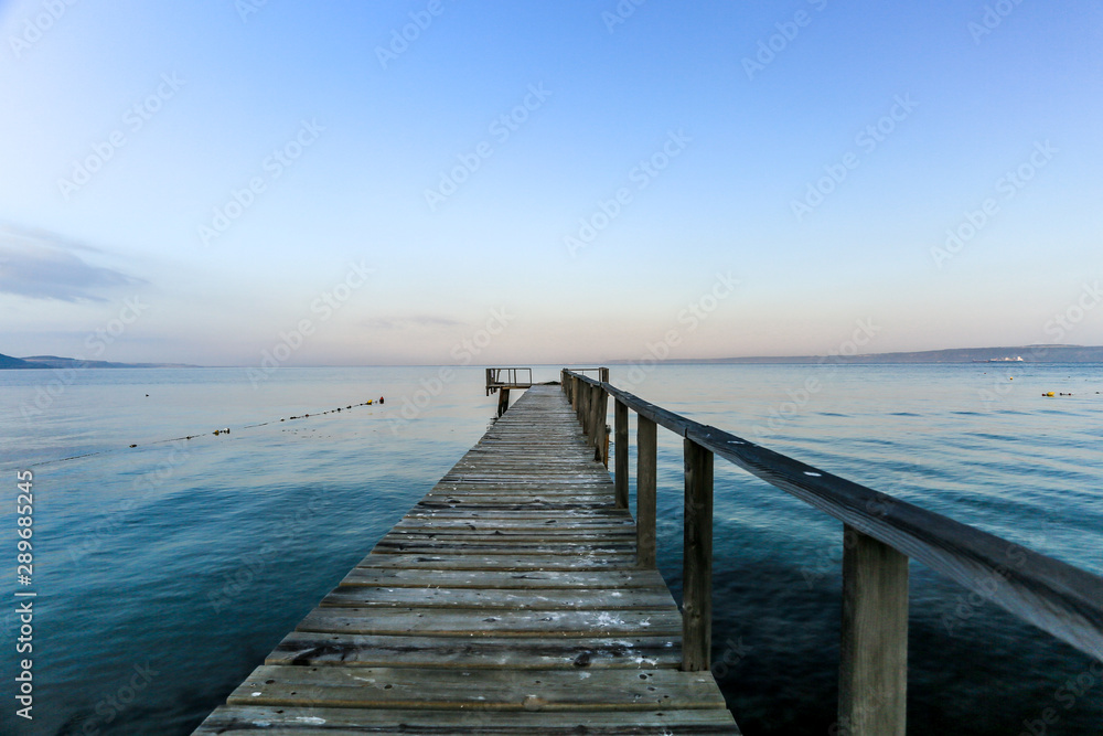A brown wood pier horizon toward to the ocean and include shade of colors, blue and orange of sky and mountain in the background also.