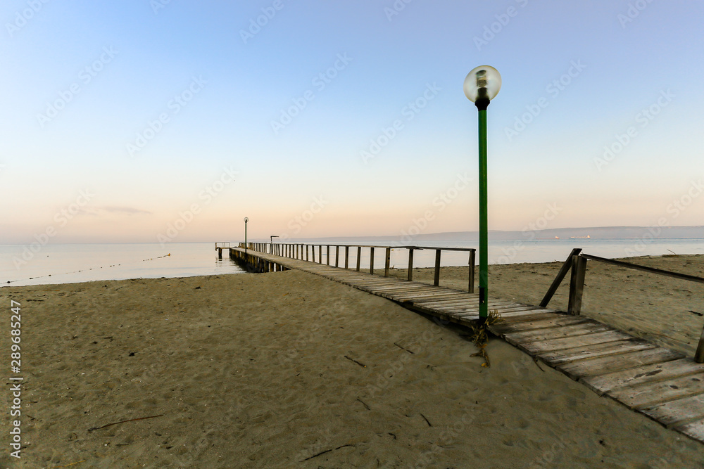 Obraz premium A brown wood pier horizon toward to the ocean that has sand beach in front a view and include shade of colors, blue and orange of sky and mountain in the background also.