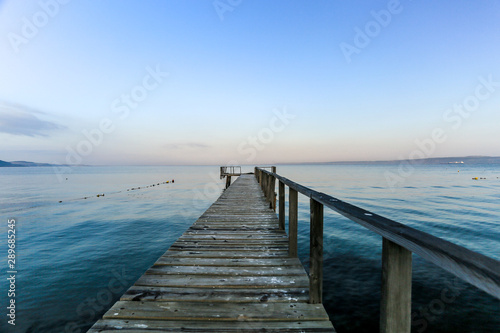 A brown wood pier horizon toward to the ocean and include shade of colors, blue and orange of sky and mountain in the background also.