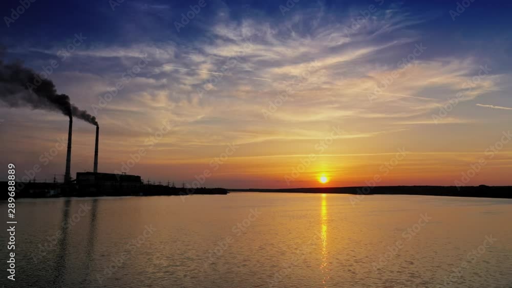 Orange sun reflects in water surface and industrial factory nearby. Beautiful evening background of setting sun on water. Manufacturing territory with smoke from pipes near the river. Orange sun reflects in water surface and industrial factory nearby. Beautiful evening background of setting sun on water. Manufacturing territory with smoke from pipes near the river.