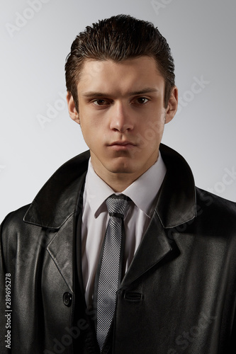 Cropped front view shot of a dark-haired man, wearing white shirt, silver spotted tie with black border and black leather jacket. He is looking straight. The photo is taking on a grey background.  photo