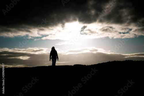 The sun disappearing behind a cloud revealing blue sky and creating a silhouette of a hiking walking off into the distance.