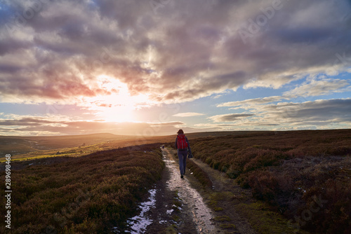 A hiker centre frame walking straight towards a sunset on open heather moors at Edmundbyers.