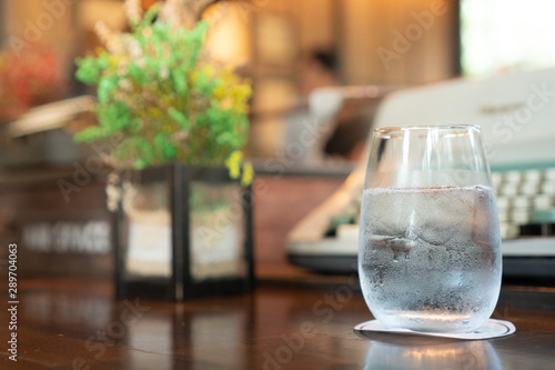 A glass of water with ice place on a dark brown wooden table with a paper coaster under a glass. there are an artificial flower and typewriter in the background © Ingon