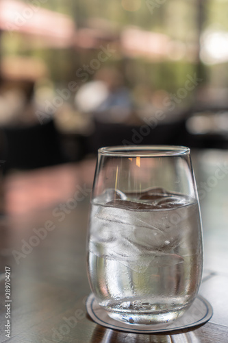 A glass of water with ice place on a dark brown wooden table with a paper coaster under a glass