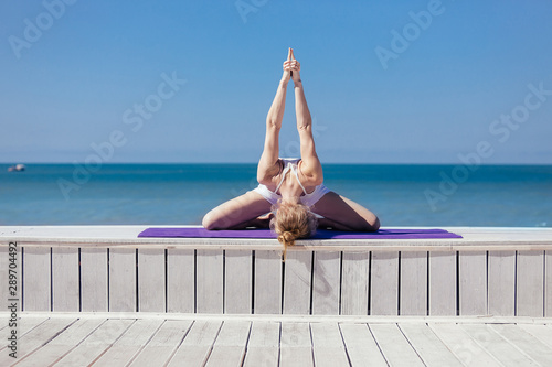 Sporty young woman sitting in variation of Upavishtha Konasana, Forward Bend posture. Slim girl practicing yoga outdoor, blue sky, white wooden terrace. Relax, healthy lifestyle concept photo