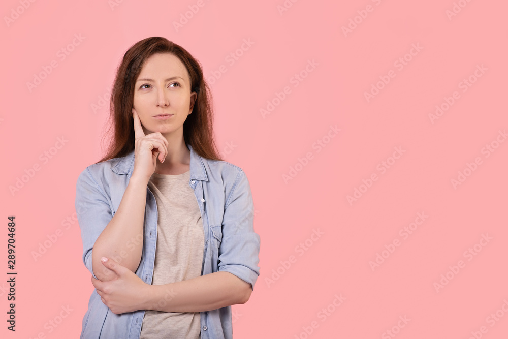 Portrait of a pensive red-haired young girl in casual clothes looking thoughtfully up posing on a pink background. Young woman chooses between goods and services. Advertising space Portrait of a pensive red-haired young girl in casual clothes looking thoughtfully up posing on a pink background. Young woman chooses between goods and services. Advertising space