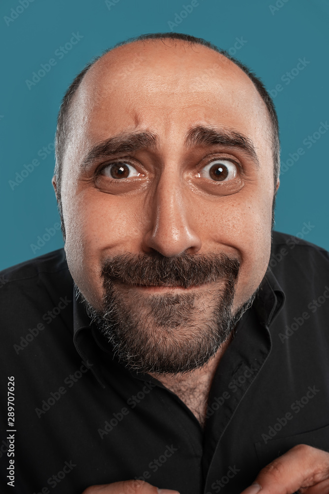 Obraz premium Close-up portrait of a brunet middle-aged man with beard, dressed in a black t-shirt and posing against a blue background.