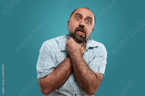 Close-up portrait of a brunet middle-aged man with beard, dressed in a light checkered shirt and posing against a blue background.