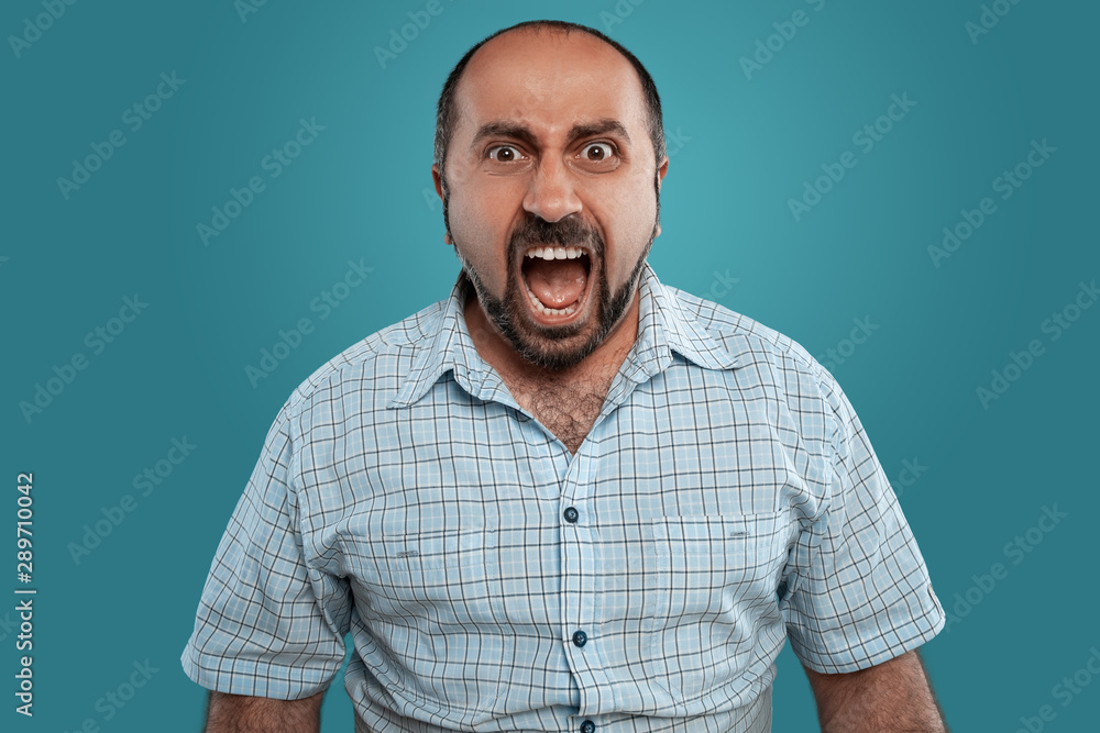 Fototapeta premium Close-up portrait of a brunet middle-aged man with beard, dressed in a light checkered shirt and posing against a blue background.