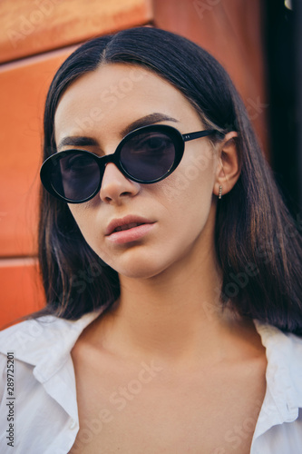 Portrait of girl in dark sunglasses posing in city against orange building. Dressed in white shirt. © nazarovsergey