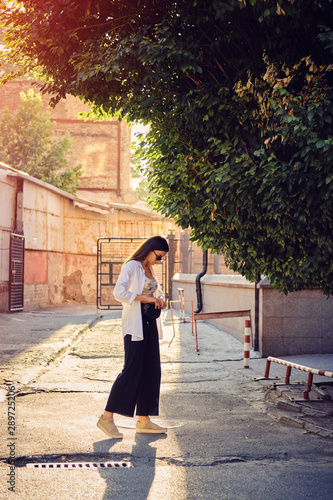 Portrait of a girl in dark sunglasses posing in city. Dressed in top with floral print, white shirt, black trousers, waist bag and sneakers.
