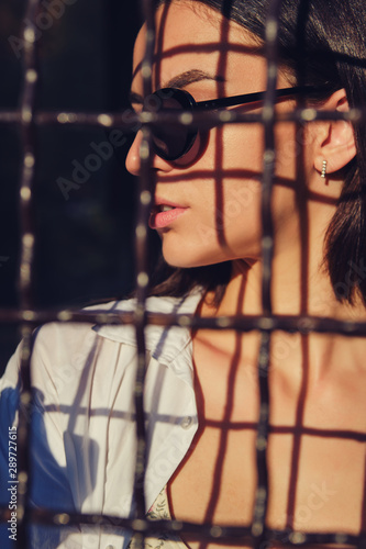 Portrait of girl in sunglasses posing in city behind a trellised fence. Dressed in top with floral print, white shirt, black trousers, waist bag.