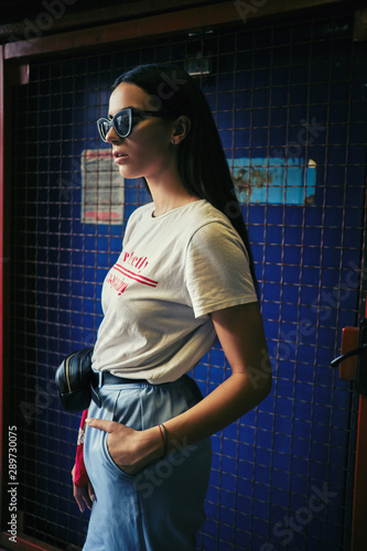 Portrait of a girl in dark sunglasses posing at a blue gate near the mall. Dressed in white t-shirt, blue trousers, black waist bag, red kerchief.