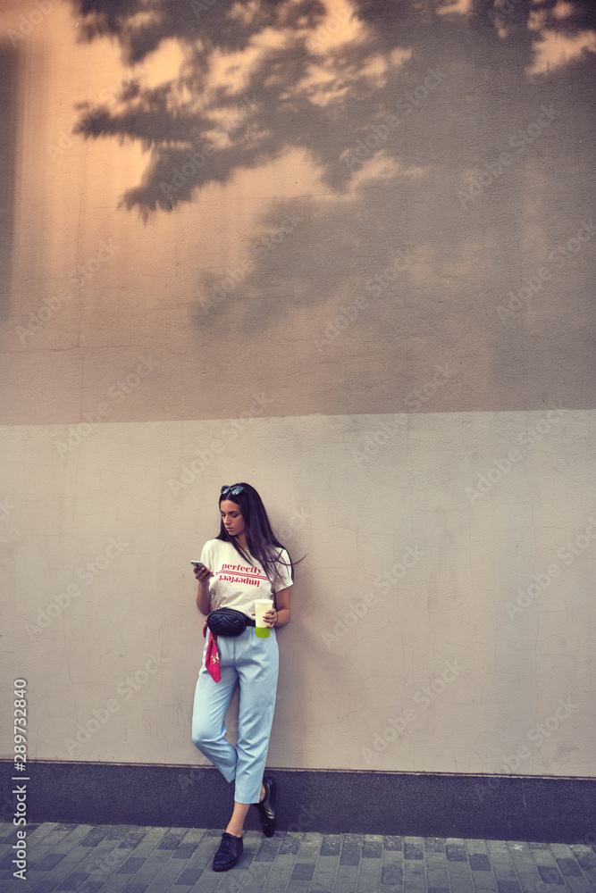 Portrait of a girl in dark sunglasses posing in city, against a wall. Dressed in white t-shirt, blue trousers, black waist bag and sneakers.