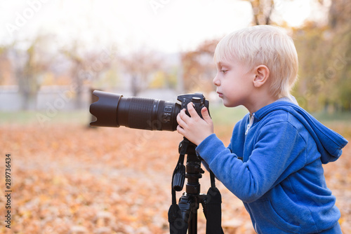 Portrait of a little blond boy with a large DSLR camera on a tripod. Photo session in the autumn park © somemeans