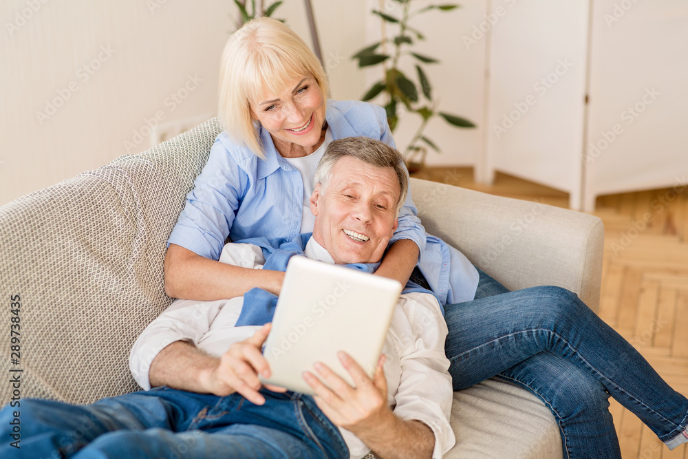 Happy mature spouses using tablet, relaxing on sofa Happy mature spouses using tablet, relaxing on sofa