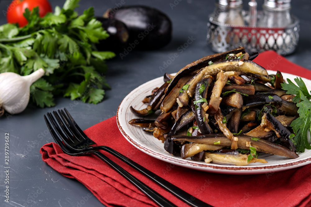 Fried eggplants with garlic and soy sauce, sliced in stripes, in a plate against a dark background Fried eggplants with garlic and soy sauce, sliced in stripes, in a plate against a dark background