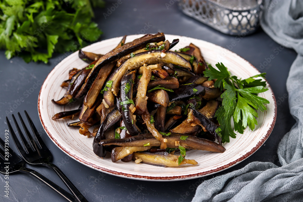 Fried eggplants with garlic and soy sauce, sliced in stripes, in a plate against a dark background
