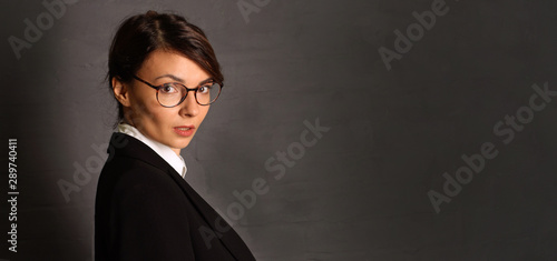 Portrait of a young brunette woman with glasses in a black suit on a dark background.
