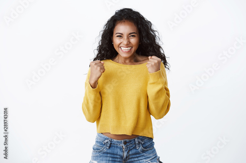 Yes deal mine. Successful african-american cute woman in yellow sweater, smiling happily, celebrating amazing news, making fist pump reacting lucky excellent opportunity, standing white background