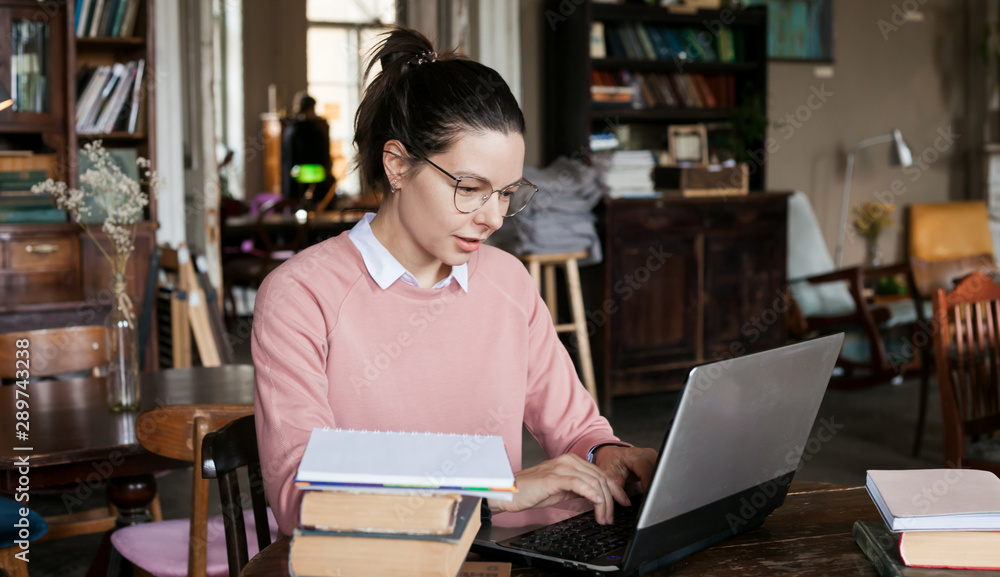 Exam preparation. Attractive brunette student girl in glasses and a pink sweater, with a slight smile sits at a laptop. Exam preparation. Attractive brunette student girl in glasses and a pink sweater, with a slight smile sits at a laptop.