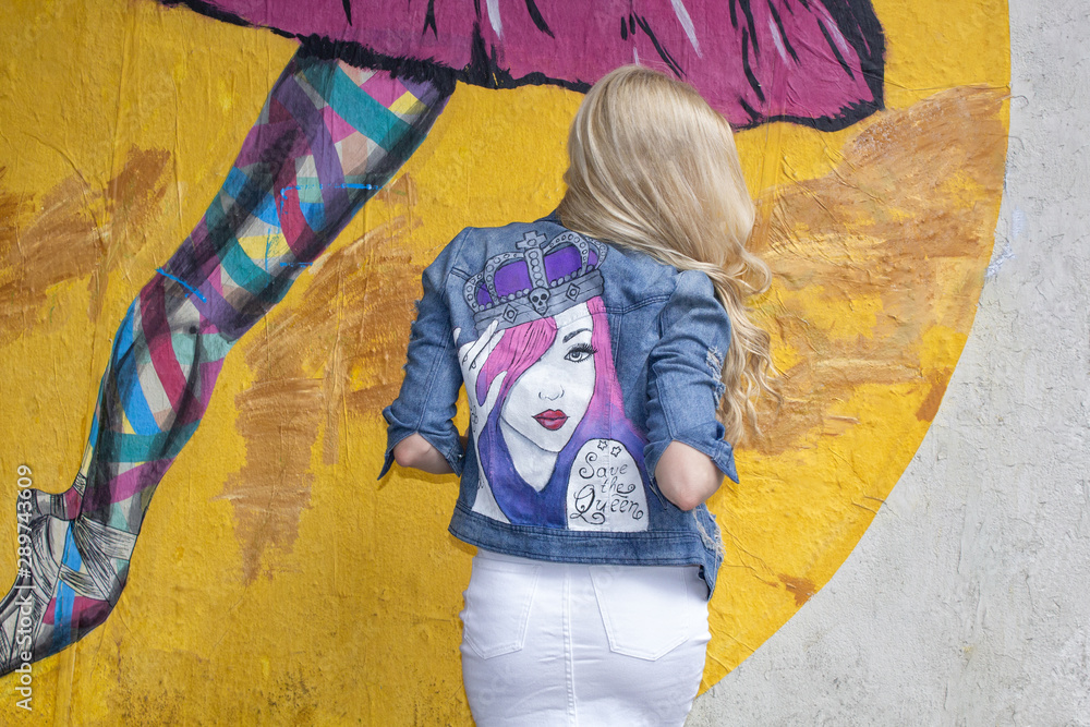 Fototapeta premium Blondynka pozuje w pomalowanej dżinsowej kurtce na tle ściany. Na ulicy.