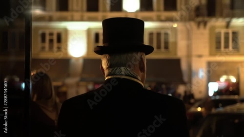 Elegant doorman at the entrance of fancy hotel. Uniformed porter meets guests at private luxury party. Doorman in elegant black suit, top hat and scarf opens the restaurant door and welcome visitors.