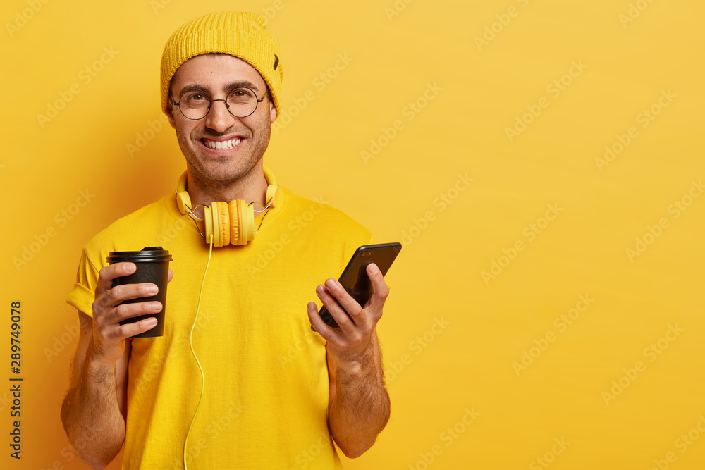Portrait of handsome cheerful young man with pleased face expression, holds mobile phone, sends text messages to friends, drinks takeaway coffee, wears spectacles, yellow outfit with headphones Portrait of handsome cheerful young man with pleased face expression, holds mobile phone, sends text messages to friends, drinks takeaway coffee, wears spectacles, yellow outfit with headphones