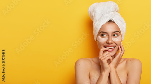 Photo of lovely young woman with fresh healthy skin, looks away, has beauty procedures, stands bare shoulders against yellow background, cares about her body, nourishes skin wears patches for softness