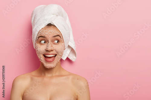Portrait of happy female model applies sea salt scrub on face, has positive expression, looks aside, has naked body, wears towel after bath, poses over pink wall with copy space, uses beauty product