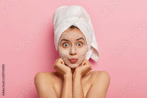 Fotografie, Obraz Horizontal shot of surprised Caucasian woman keeps palms under chin, looks with widely eyes, applies scrub mask, avoids problems with skin, stands shirtless against pink background