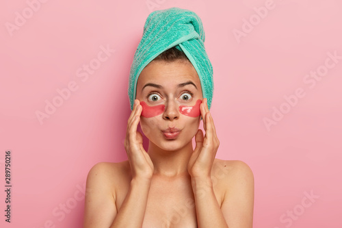 Headshot of shocked bare shoulders woman has natural beauty, reduces wrinkles under eyes, applies collagen patches, has wrapped towel on head, isolated over pink background. Cosmetic treatment concept