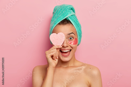 Portrait of overjoyed funny woman keeps cosmetic sponge on eye, laughs sincerely, wears undereye patches, removes wrinkles, cares about skin, has natural beauty, isolated over pink background