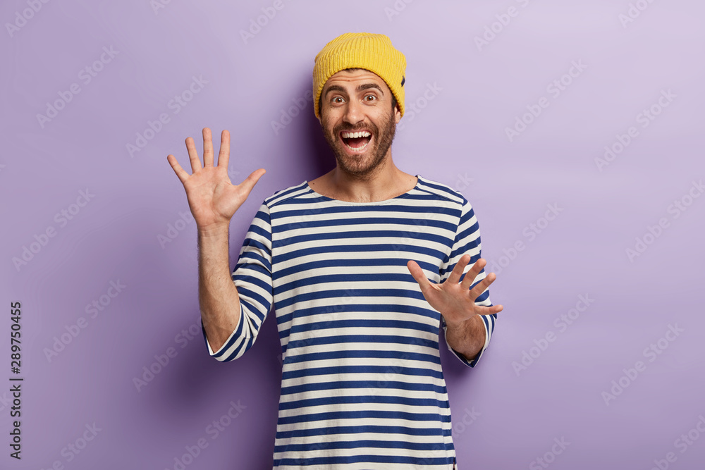 Overjoyed unshaven millennial guy gestures actively, shows palms at camera, being in good mood, wears yellow hat and striped jumper, isolated over purple background. People, emotions, happiness Overjoyed unshaven millennial guy gestures actively, shows palms at camera, being in good mood, wears yellow hat and striped jumper, isolated over purple background. People, emotions, happiness