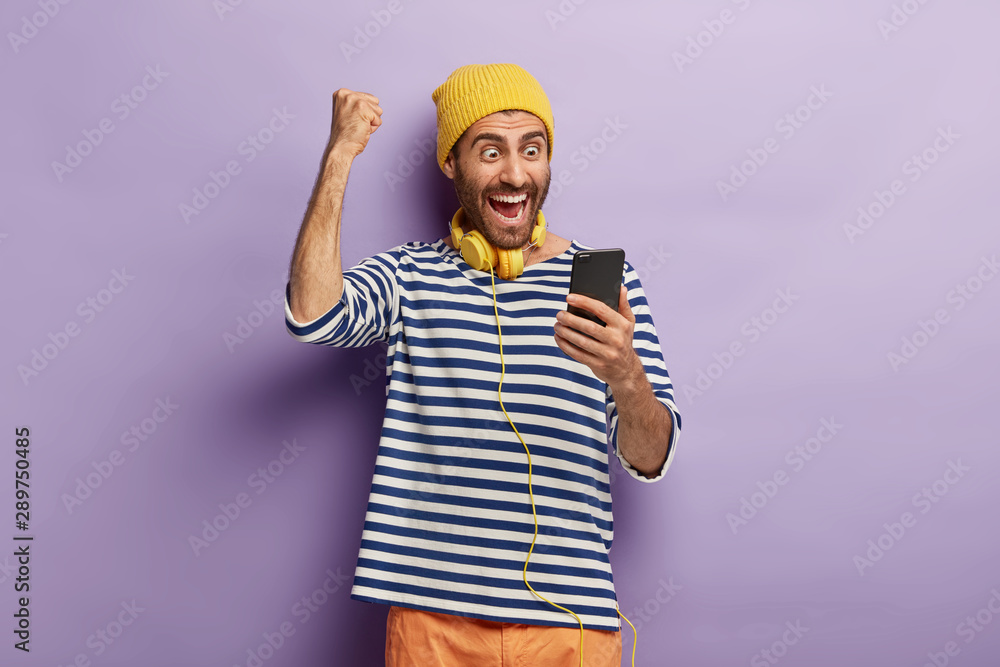 Triumphing happy guy raises clenched fist, celebrates winning lottery, gets message of confirming holds mobile phone, browses social media, wears yellow hat, striped jumper, always stays in touch Triumphing happy guy raises clenched fist, celebrates winning lottery, gets message of confirming holds mobile phone, browses social media, wears yellow hat, striped jumper, always stays in touch