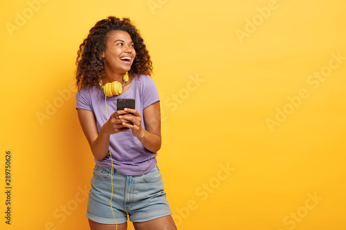 Photo of smiling teenage girl with Afro haircut, uses smartphone for listening music in playlist, wears headphones, looks positively aside, dressed in everyday summer clothes copy space on yellow wall