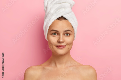 Good looking young woman with smooth healthy skin, has naked body, looks straightly at camera, has blue eyes, wears towel on head, takes shower in bathroom, has hygienic treatments every morning