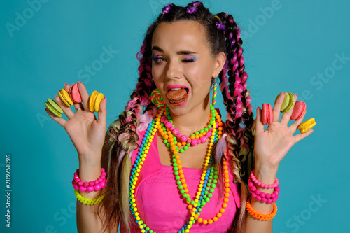 Lovely girl with multi-colored braids hairstyle and bright make-up  holding macarons between her fingers  posing in studio against a blue background.