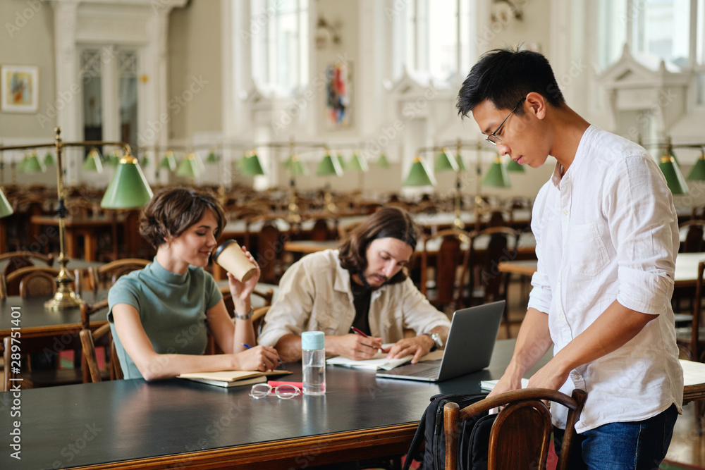 Fototapeta premium Young asian student in eyeglasses with backpack coming to study with friends in library of university