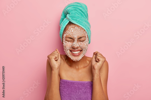 Energized happy dark skinned female model raises clenched fists, rejoices pleasant moment in life, has eyes closed, toothy smile, applies sea salt srcub mask on complexion for reducing fine lines photo