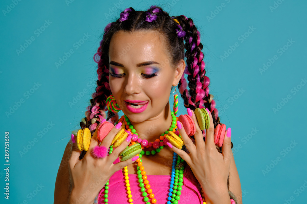 Lovely girl with multi-colored braids hairstyle and bright make-up, holding macarons between her fingers, posing in studio against a blue background. Lovely girl with multi-colored braids hairstyle and bright make-up, holding macarons between her fingers, posing in studio against a blue background.