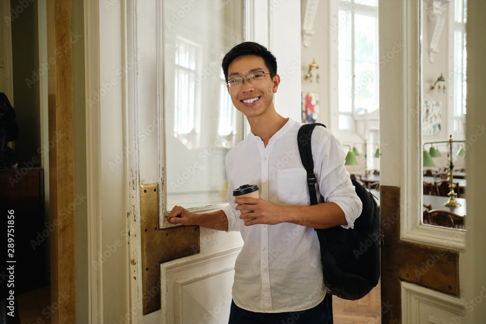 Young asian male student in eyeglasses with backpack and coffee to go opening library door in university Young asian male student in eyeglasses with backpack and coffee to go opening library door in university