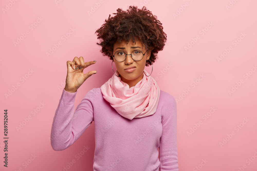 Upset curly haired woman shapes little gesture, demonstrates something very tiny, wears optical glasses, casual jumper and scarf, measures small size, isolated over pink background. Not very much Upset curly haired woman shapes little gesture, demonstrates something very tiny, wears optical glasses, casual jumper and scarf, measures small size, isolated over pink background. Not very much