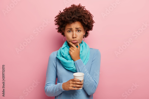 Photo of frustrated unhappy woman holds takeaway coffee, tries to solve problem, being overworked, holds chin and looks sadly at camera, wears blue neat clothes, poses against rosy studio wall