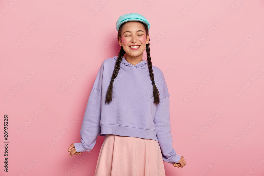 Positive feelings concept. Satisfied adorable young Korean female model with rouge cheeks, laughs from joy, wears blue cap, oversized jumper and skirt, isolated on pink, expresses good emotions Positive feelings concept. Satisfied adorable young Korean female model with rouge cheeks, laughs from joy, wears blue cap, oversized jumper and skirt, isolated on pink, expresses good emotions