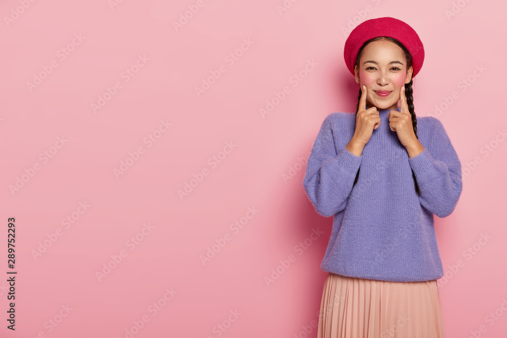 Positive young woman keeps fore fingers on both cheeks, being in high spirit, wears red beret, purple sweater and skirt, stands over pink wall, copy space aside, shows her beauty and vivid makeup Positive young woman keeps fore fingers on both cheeks, being in high spirit, wears red beret, purple sweater and skirt, stands over pink wall, copy space aside, shows her beauty and vivid makeup