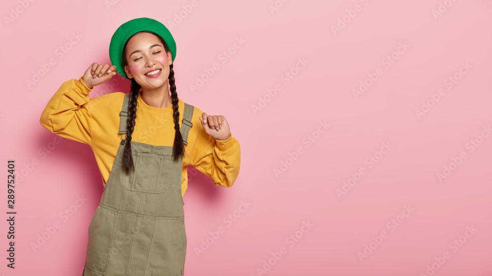Studio shot of cheerful young Asian woman spreads arms sideways, feels carefree and relaxed, enjoys nice time, has two pigtails, dressed in fashionable outfit, isolated on pink wall, blank space aside Studio shot of cheerful young Asian woman spreads arms sideways, feels carefree and relaxed, enjoys nice time, has two pigtails, dressed in fashionable outfit, isolated on pink wall, blank space aside