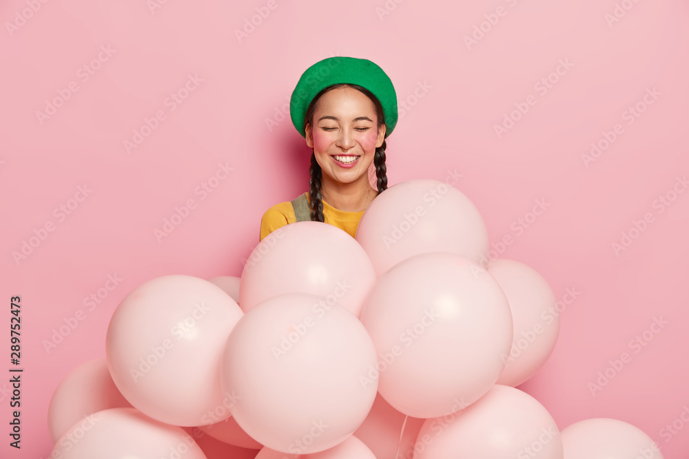 Smiling woman with cheerful expression, keeps eyes closed from pleasure, wears green beret, stands with inflated helium balloons, has two pigtails, gets congratulation with birthday or anniversary Smiling woman with cheerful expression, keeps eyes closed from pleasure, wears green beret, stands with inflated helium balloons, has two pigtails, gets congratulation with birthday or anniversary
